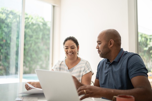 Two people sit at a table looking at a laptop and paperwork.