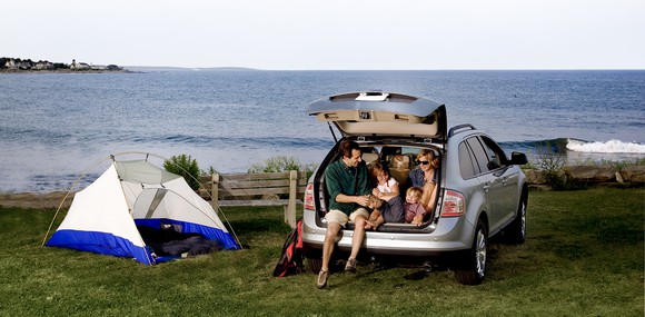 A family sitting in the back of their SUV while camping on a hill overlooking the ocean.
