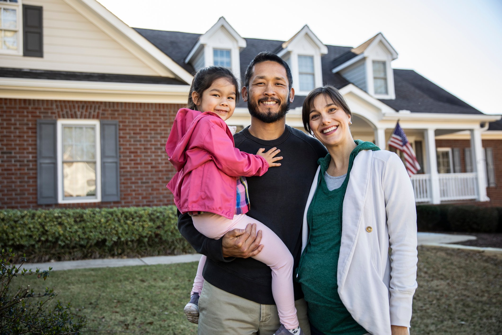 Family of three smiling and standing outside in front of a house.