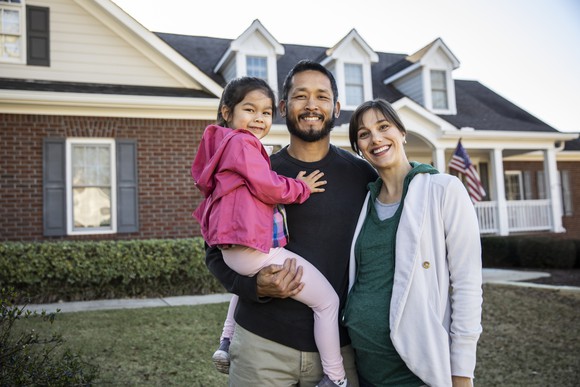 Family of three smiling and standing outside in front of a house.