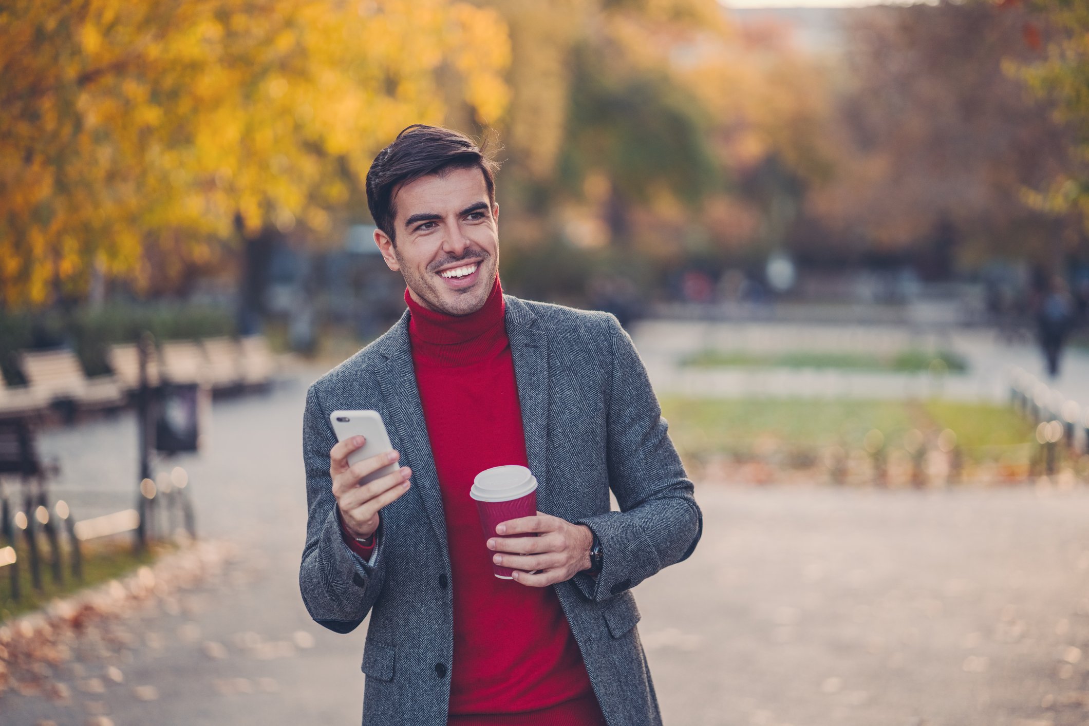 Smiling person with phone and coffee cup walking through park in the fall. 