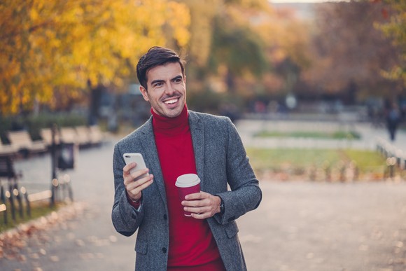 Smiling person with phone and coffee cup walking through park in the fall. 