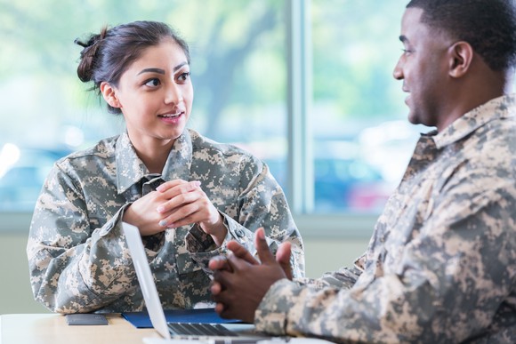 Two soldiers chat while working on a laptop.