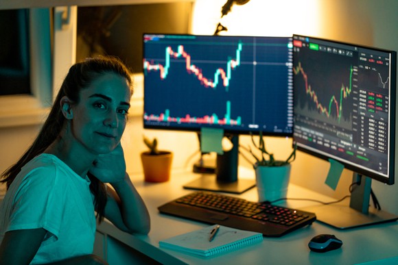A person sits in front of two computer monitors showing stock performance.