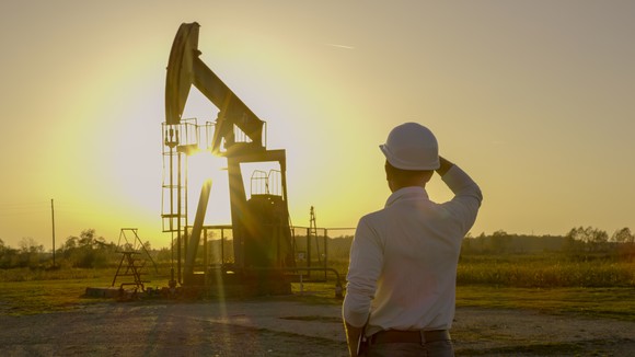A worker in a hard hat looks at an oil pumpjack with a glowing sun in the background. 