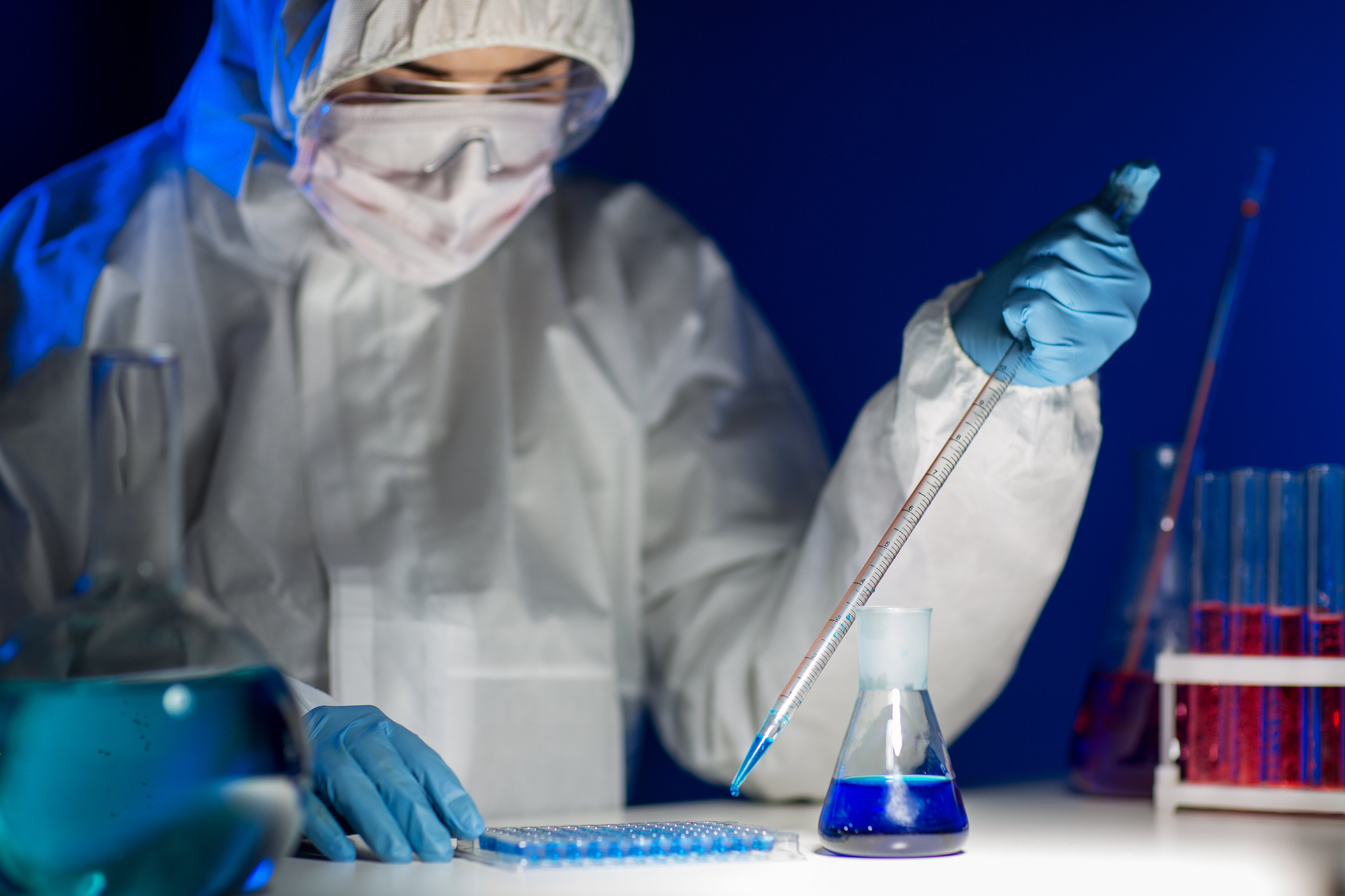 A lab technician in protective equipment using a pipette to place liquid samples into a test tray.