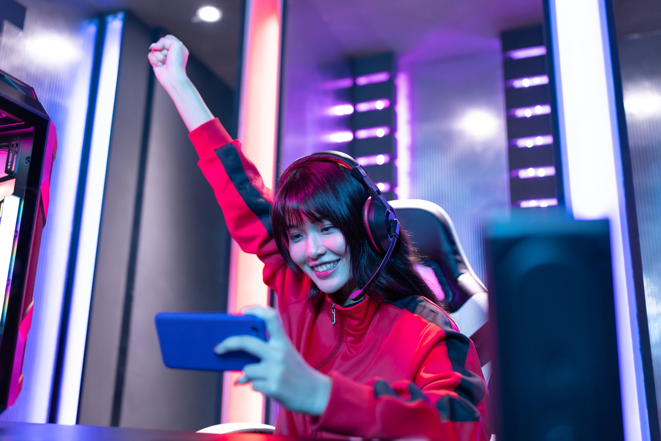 A woman pumping her right fist in the air during a gaming tourney, while holding a smartphone in her left hand.