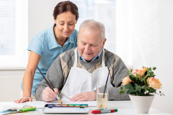 A person paints at a table with flowers on it while a caregiver watches over their shoulder.
