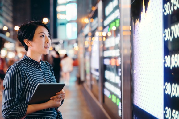 A woman holding a tablet computer stands on a sidewalk and looks at a large monitor showing stock information.
