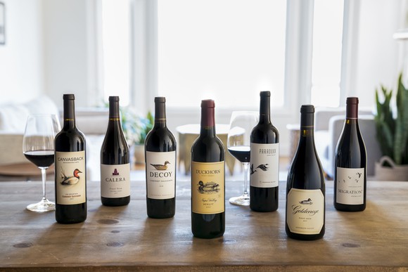 A lineup of wine bottles showing seven of Duckhorn's brands, on a wooden table in front of a bright window. 