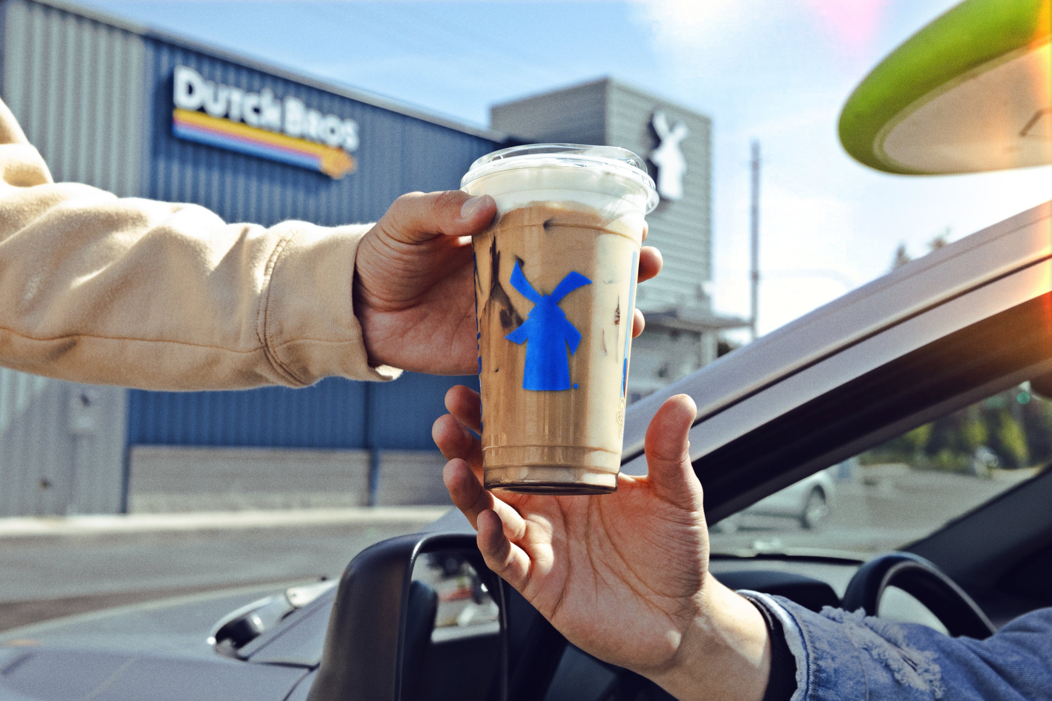 A Dutch Bros employee handing a customer a cold brew with the windmill logo at a drive-thru, with the Dutch Bros logo on the wall in the background.