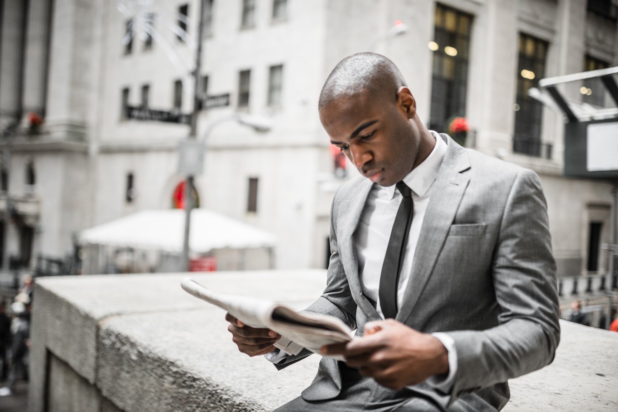 A man in a suit reads a newspaper in a city setting.
