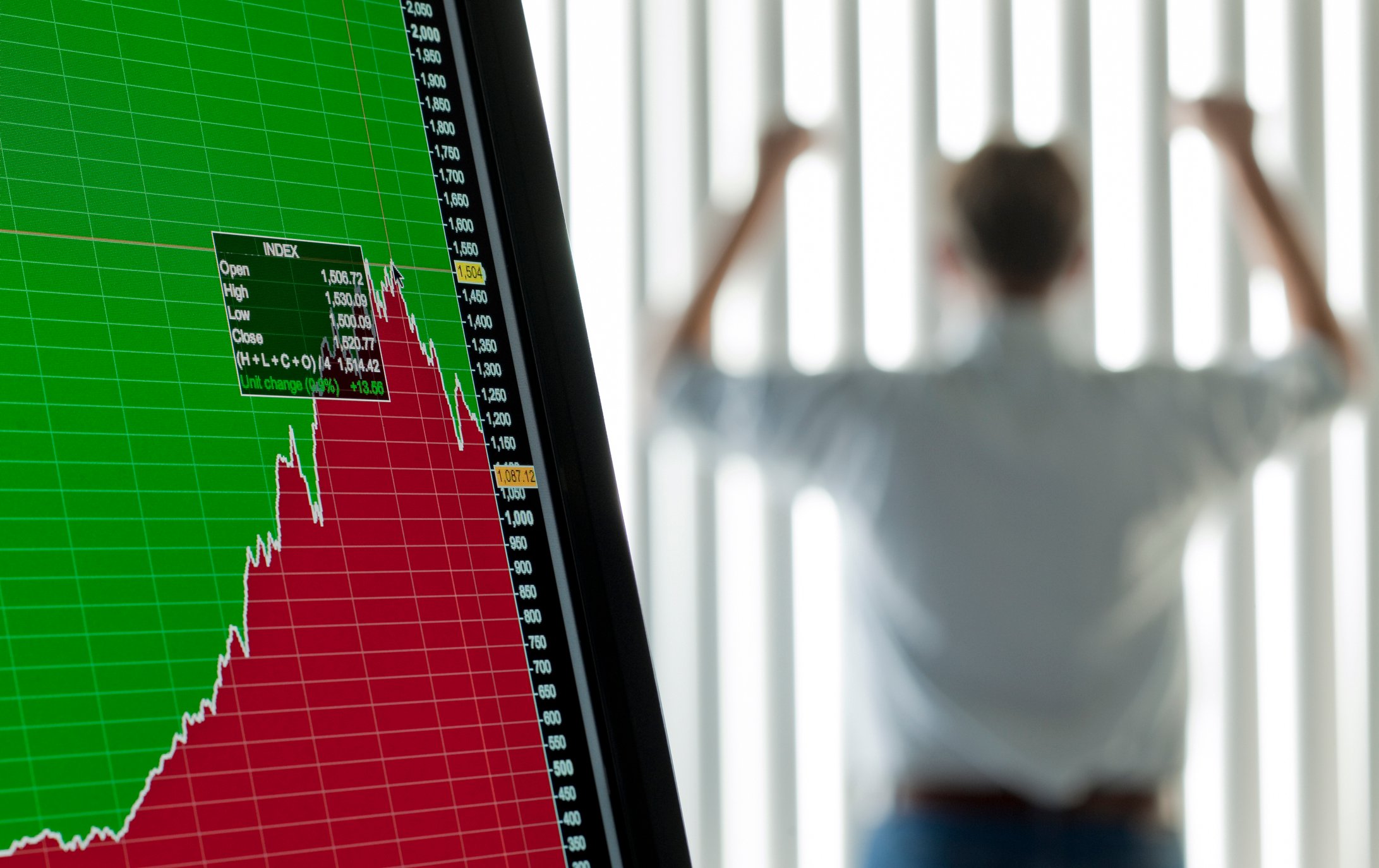 A man holds up his arms in celebration in the background as a fever chart on a computer screen in the foreground shows a downturn in value.