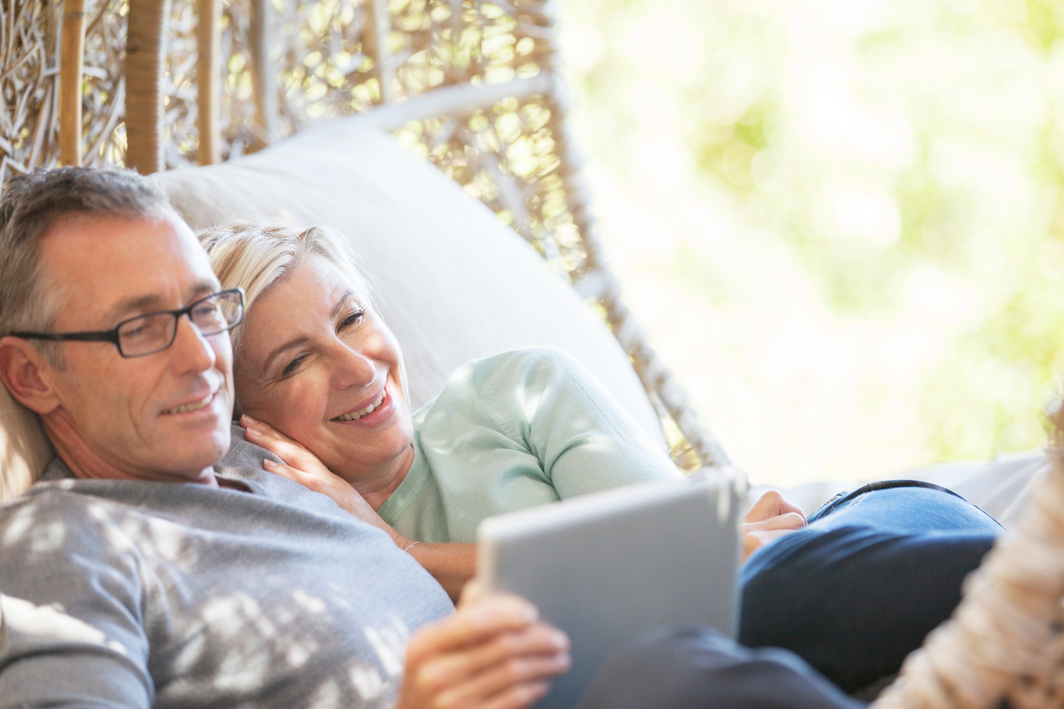 Two people lean back on cushions in a shady outdoor area and smile as they look at a tablet.