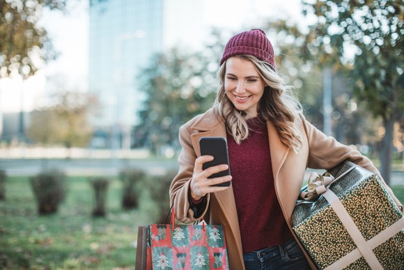 A woman smiles as she looks at her phone and carries packages through a park.