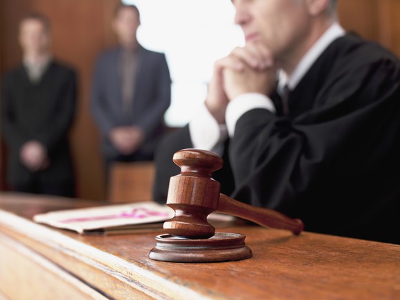A judge sitting at the bench next to a gavel with two people in the background.