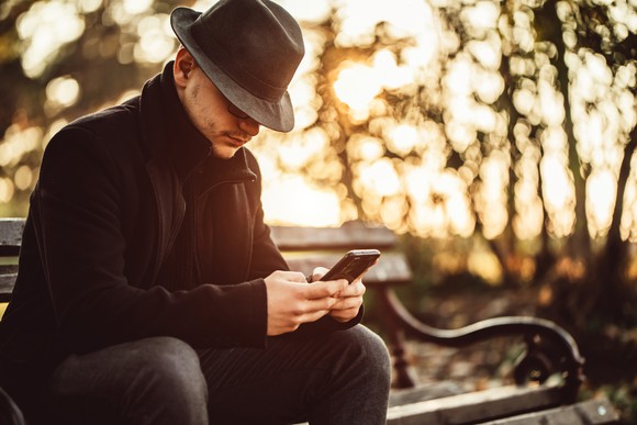 A stereotypical secret agent type with fedora pulled down low, collar turned up, texting on his phone while sitting on a bench. 