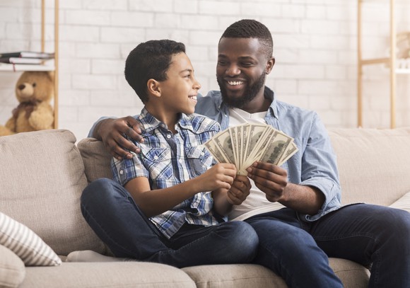 A smiling adult and child sitting on a couch holding several fanned out dollar bills.