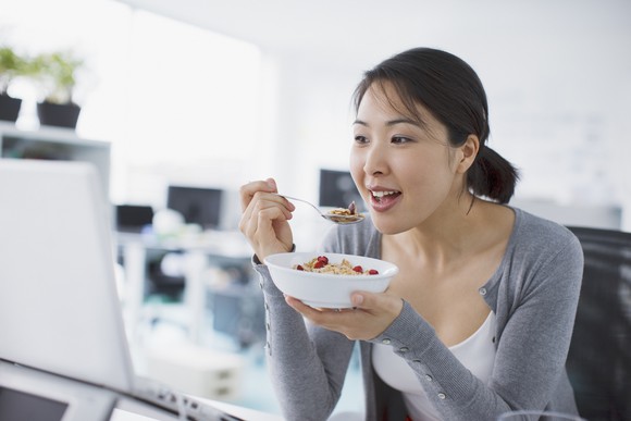 Young woman eats cereal from a bowl while looking at her computer. 
