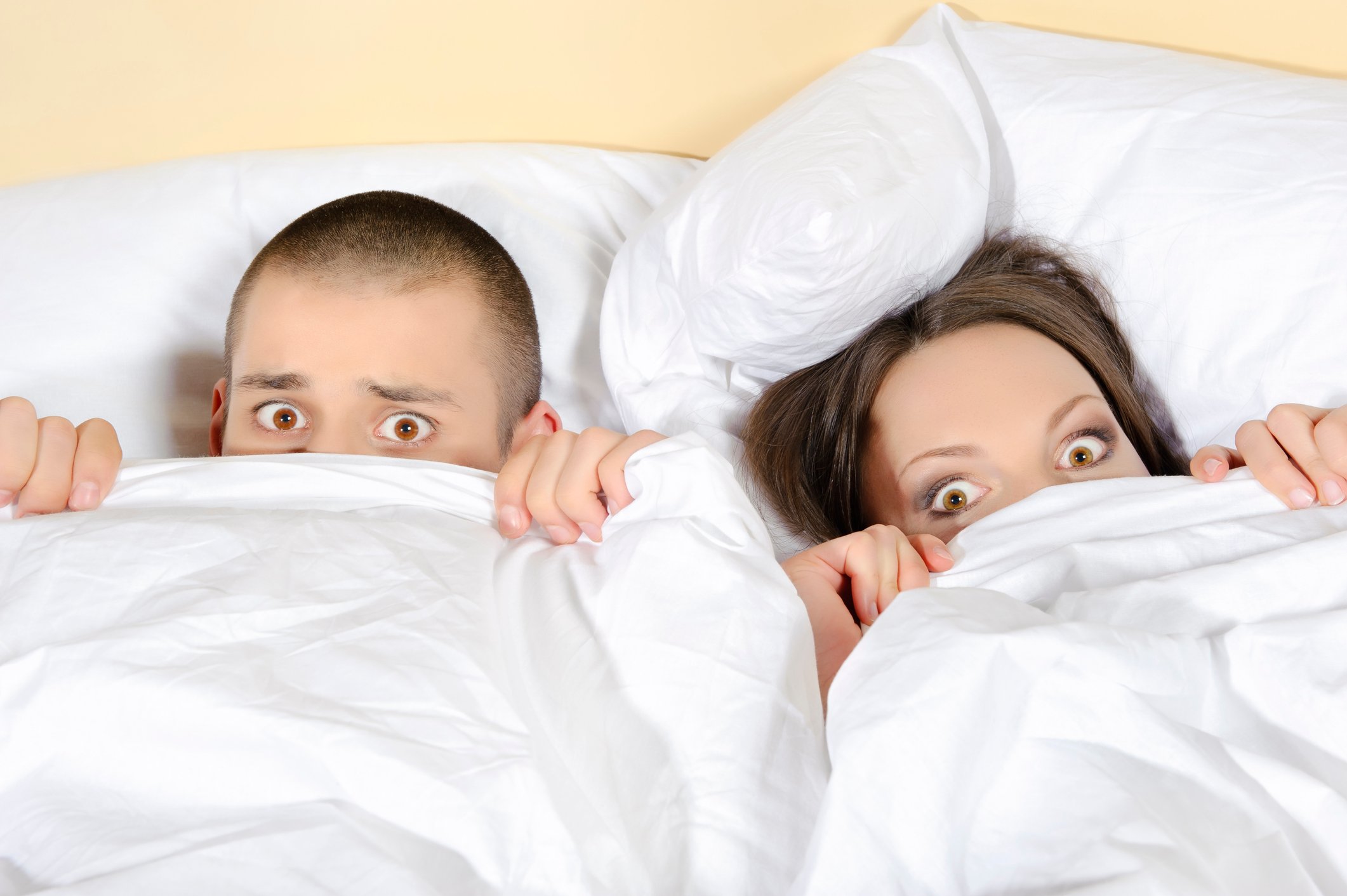 Two people in bed covering the lower halves of their faces with a comforter.