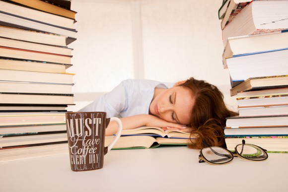 A person sleeping with their head on a book while between two stacks of books on the desk.