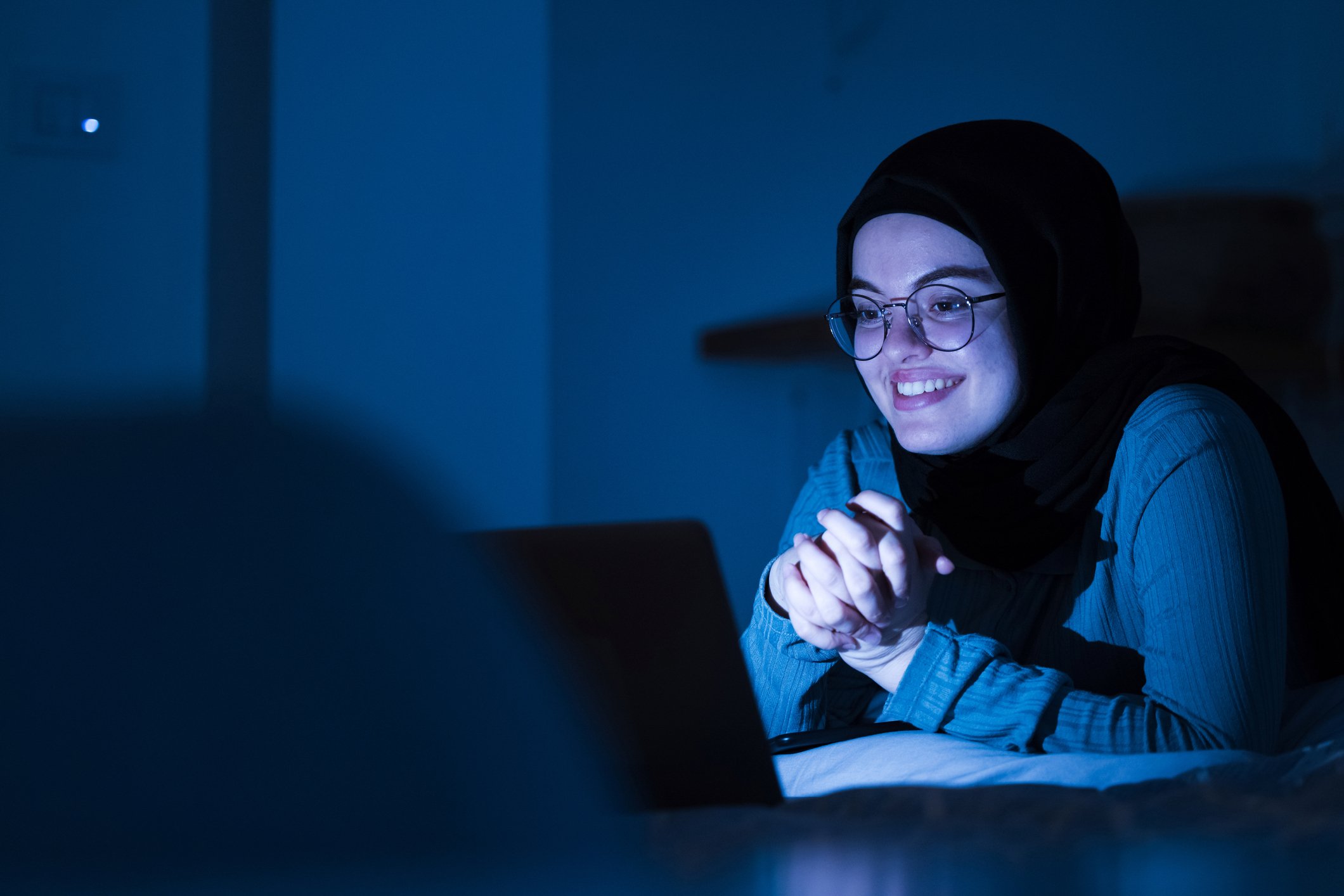 A young adult watches a movie on a laptop in a dark room.