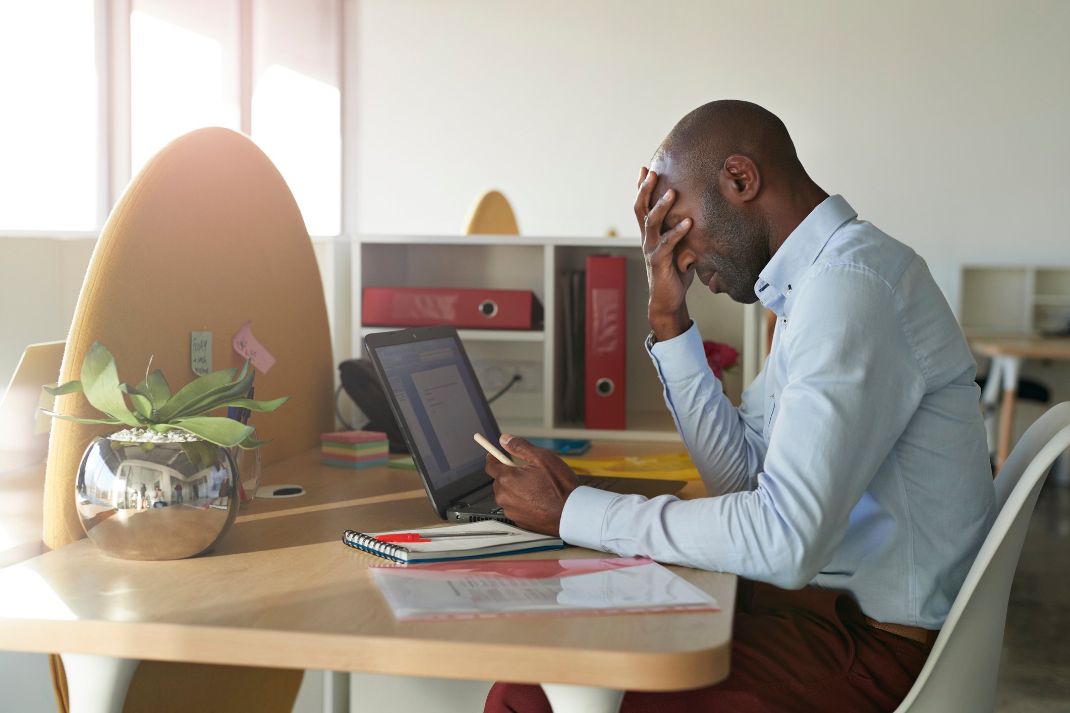 A slumped-over man at a desk covers his face with one hand while holding a phone with the other. 