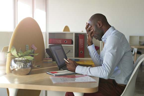 A slumped-over man at a desk covers his face with one hand while holding a phone with the other. 