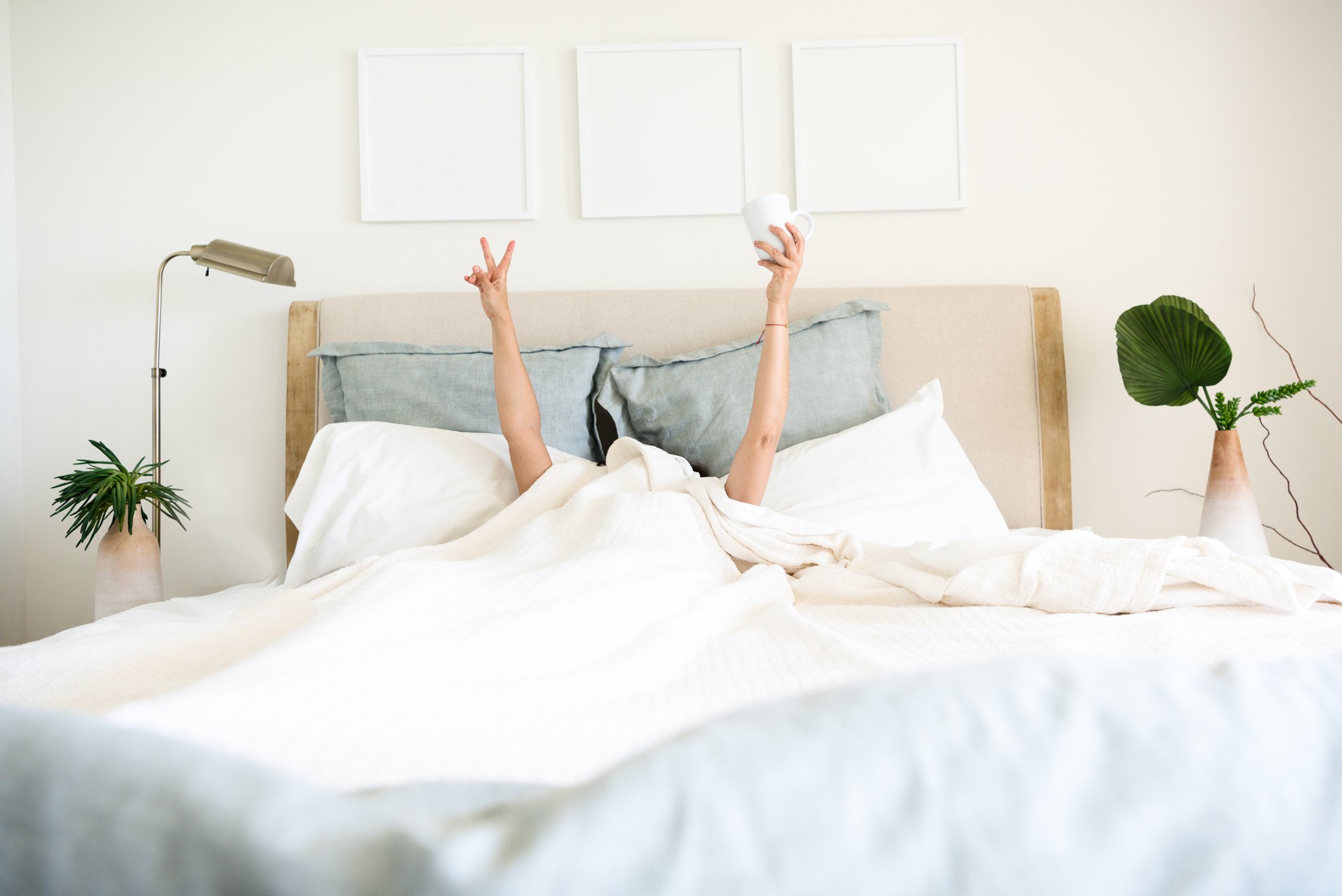 Person in bed undercovers with arms outstretched making the peace sign with one hand and holding a coffee cup in the other.