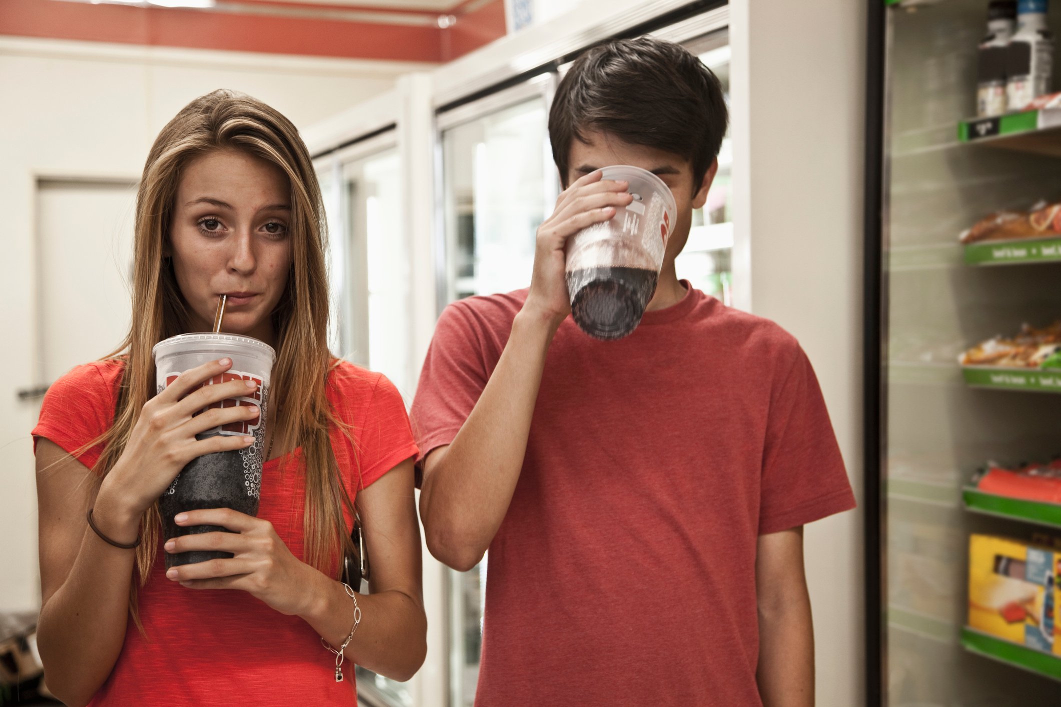 Two people holding sodas, one is drinking from the straw with a visible face and the other is sipping from the edge of the cup, their face covered.