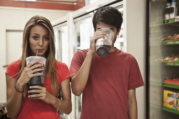 Two people holding sodas, one is drinking from the straw with a visible face and the other is sipping from the edge of the cup, their face covered.