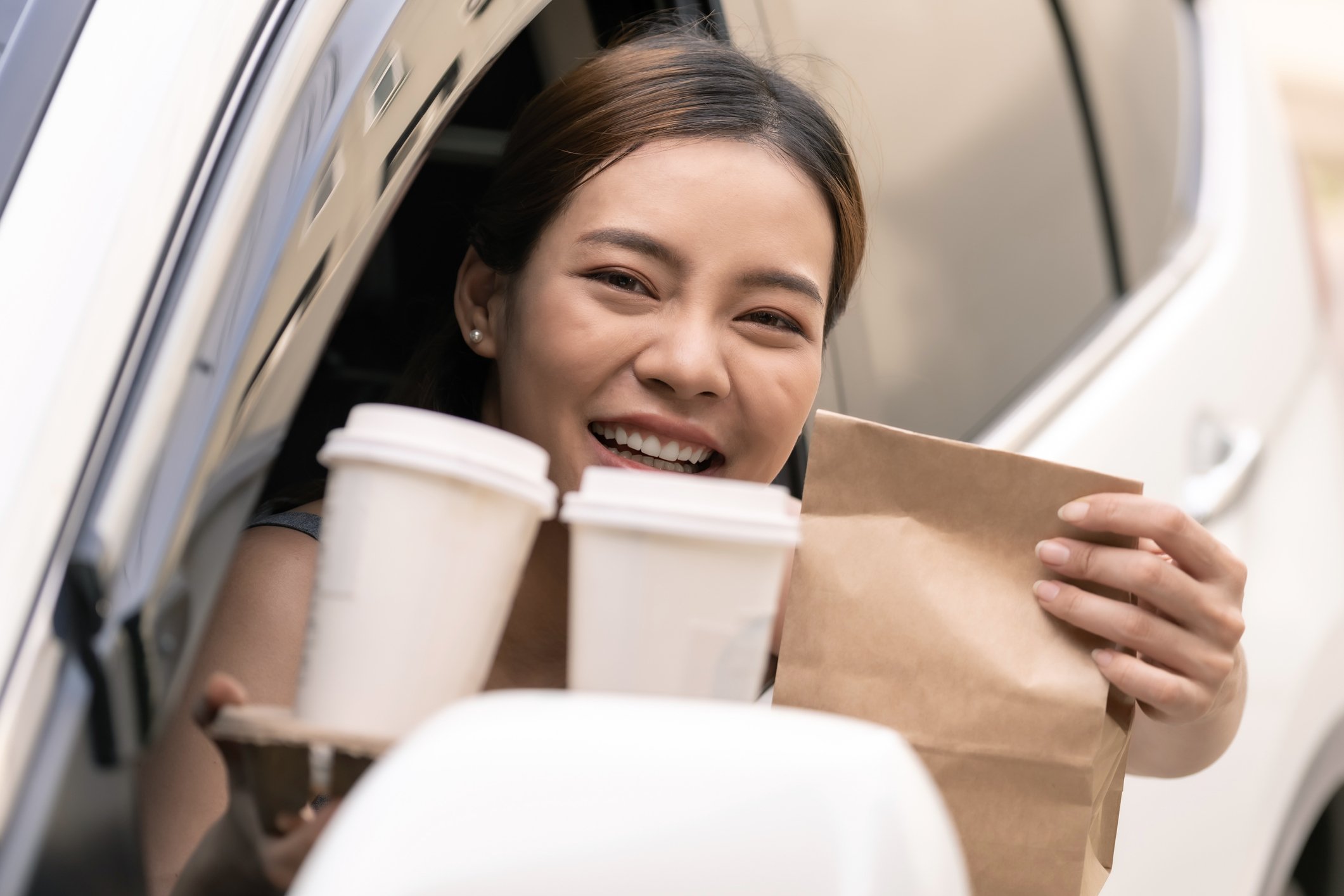 A smiling person at a drive-thru restaurant holding a bag and a cup holder with two drinks.