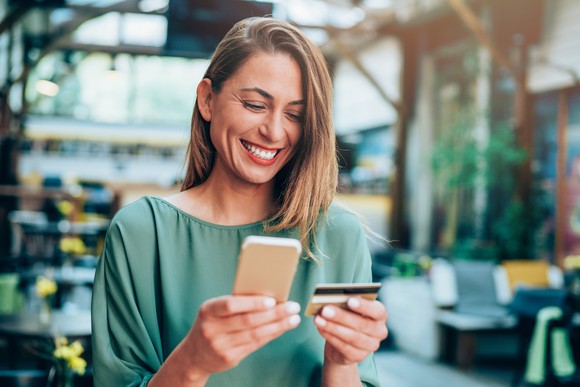 A woman looking at her phone, holding a credit card, smiling. 