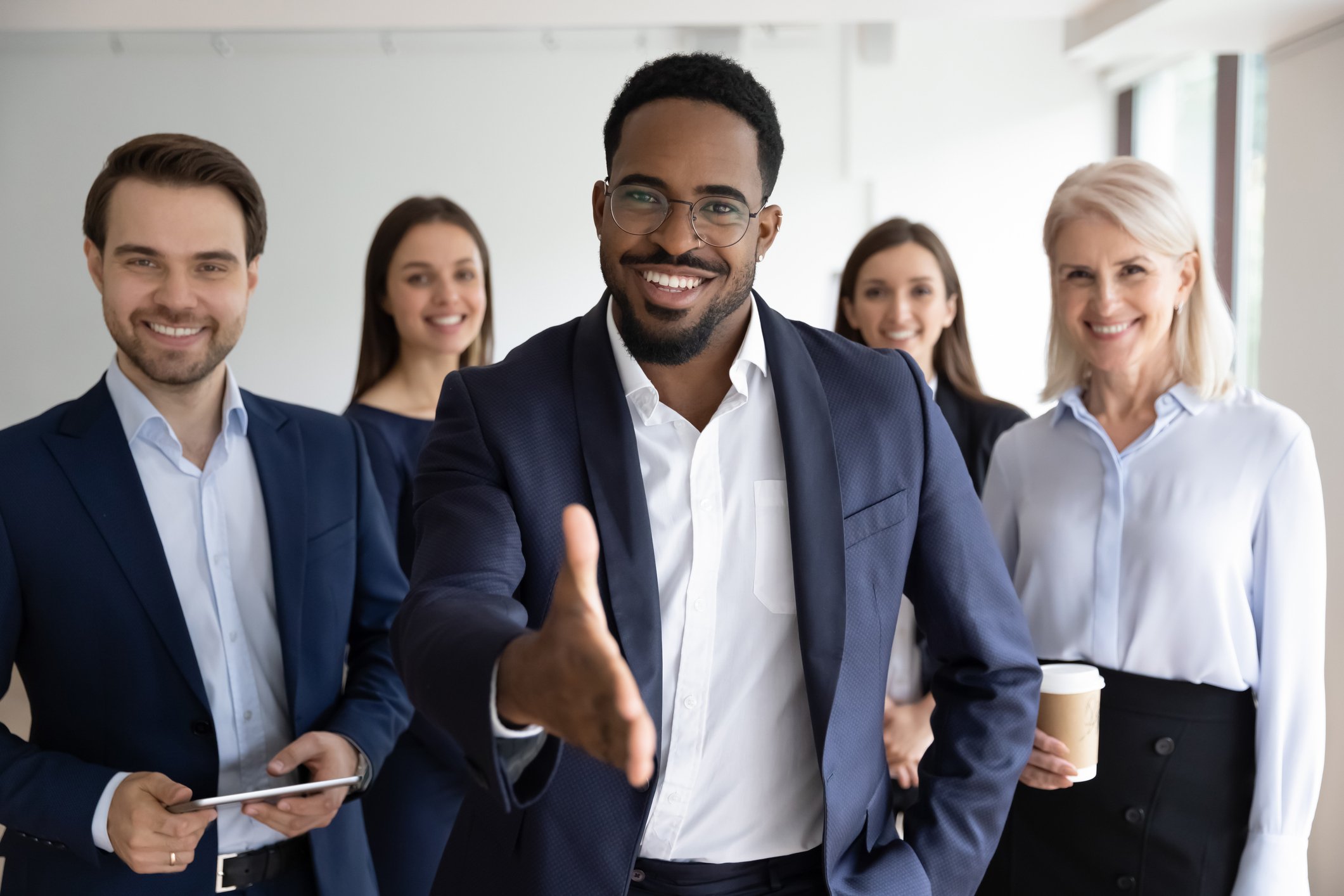 A group in business attire looking at the camera with one person reaching out to shake hands.