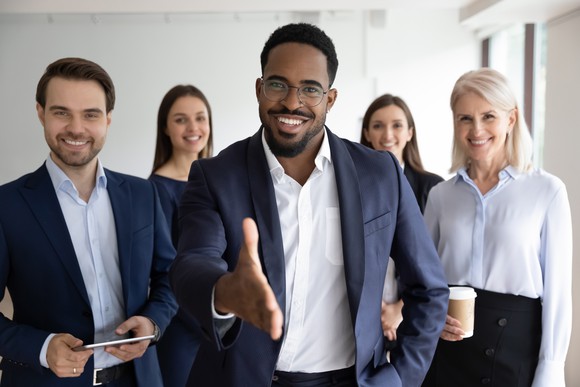 A group in business attire looking at the camera with one person reaching out to shake hands.