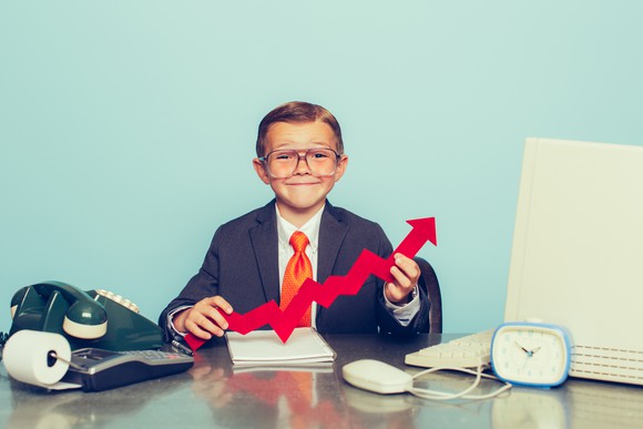 Child in suit holding stock market chart arrow at a desk.