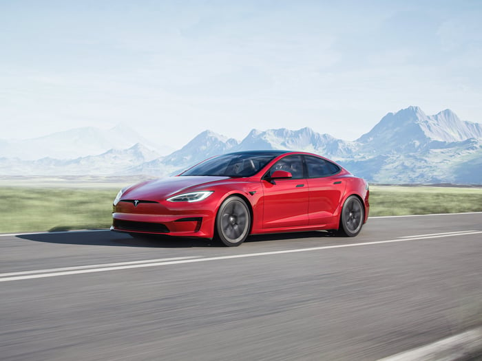 A red Tesla vehicle on a road in front of a mountain range.