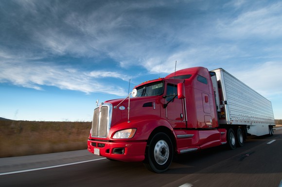 A semi with a red tractor and white trailer speeding along a highway under a partly cloudy sky. 