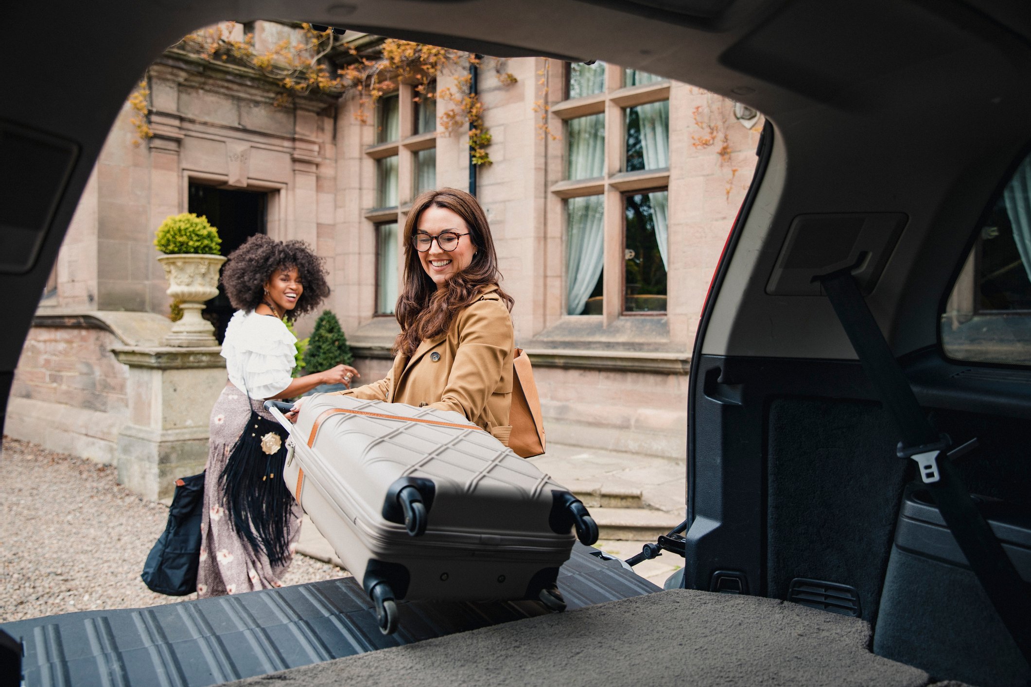 Two travelers carrying suitcases into a mansion.