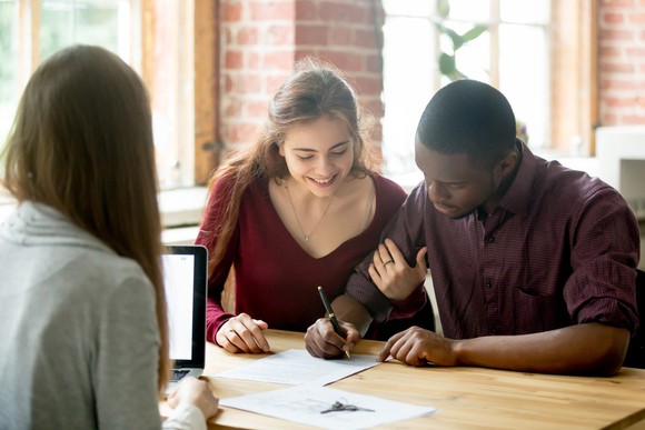 Two people signing documents at a desk in front of a third person.
