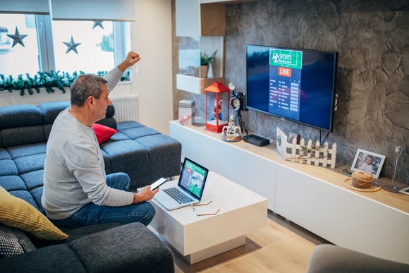 A person sitting on a couch with his hand raised looks at a sports-betting page on TV.
