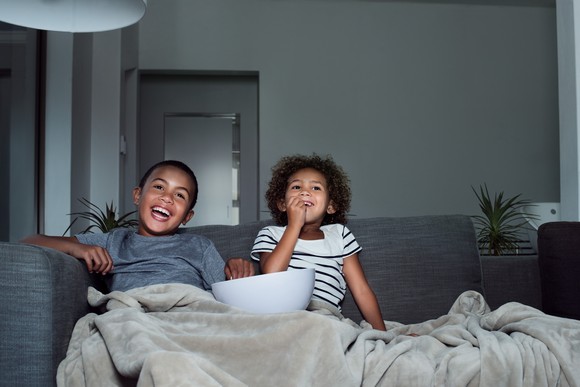 Two kids watching television on their couch and eating a snack.