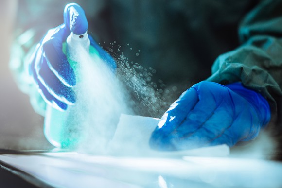 A person wearing protective clothing and blue latex gloves spraying and scrubbing a surface with disinfectant.