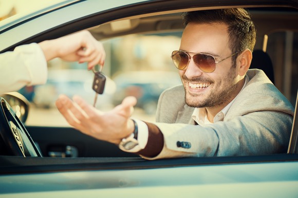 A smiling person sitting inside a new car, being handed the keys by another person. 