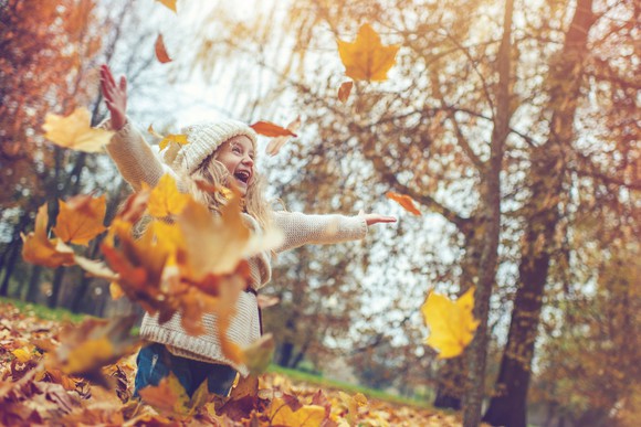 A child smiles with outstretched arms while playing in a pile of bright orange fall leaves. 