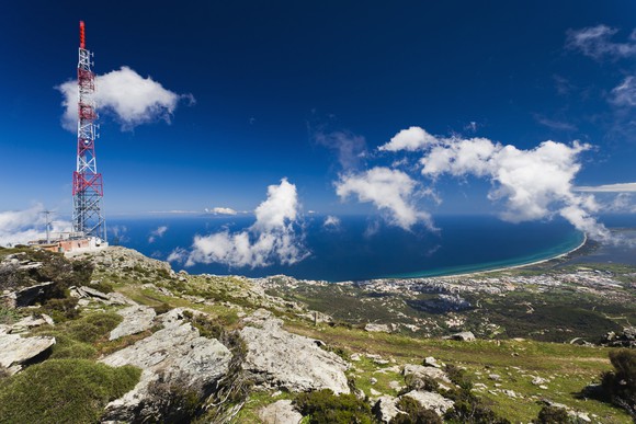 Cell phone tower on a rocky hillside against a bright blue sky with clouds. 