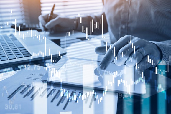 A persons hands. One is holding a pencil near paper, the other is touching a tablet that is resting on top of financial documents.