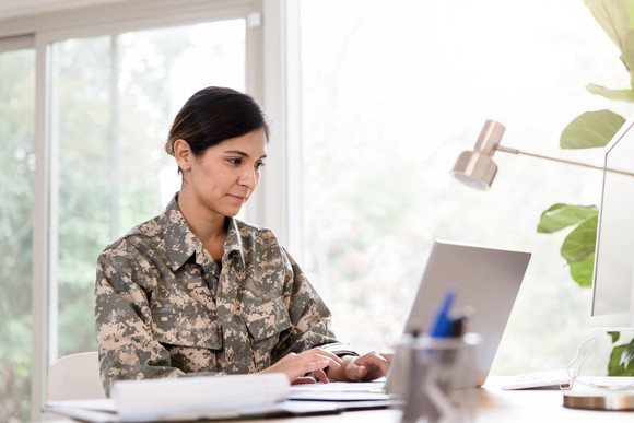 A military person is sitting at a desk, in front of an open laptop.