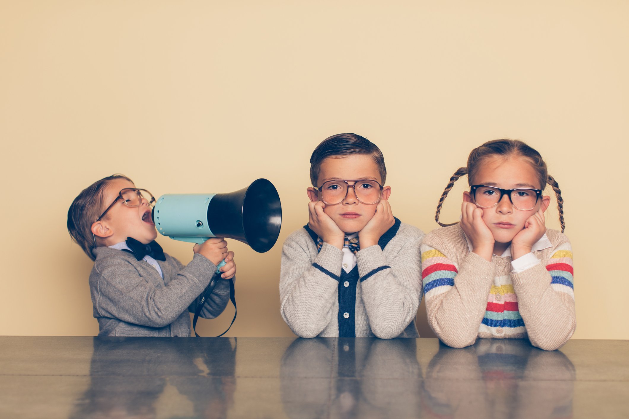 A child holds a megaphone directed at two other children with their hands over their ears.