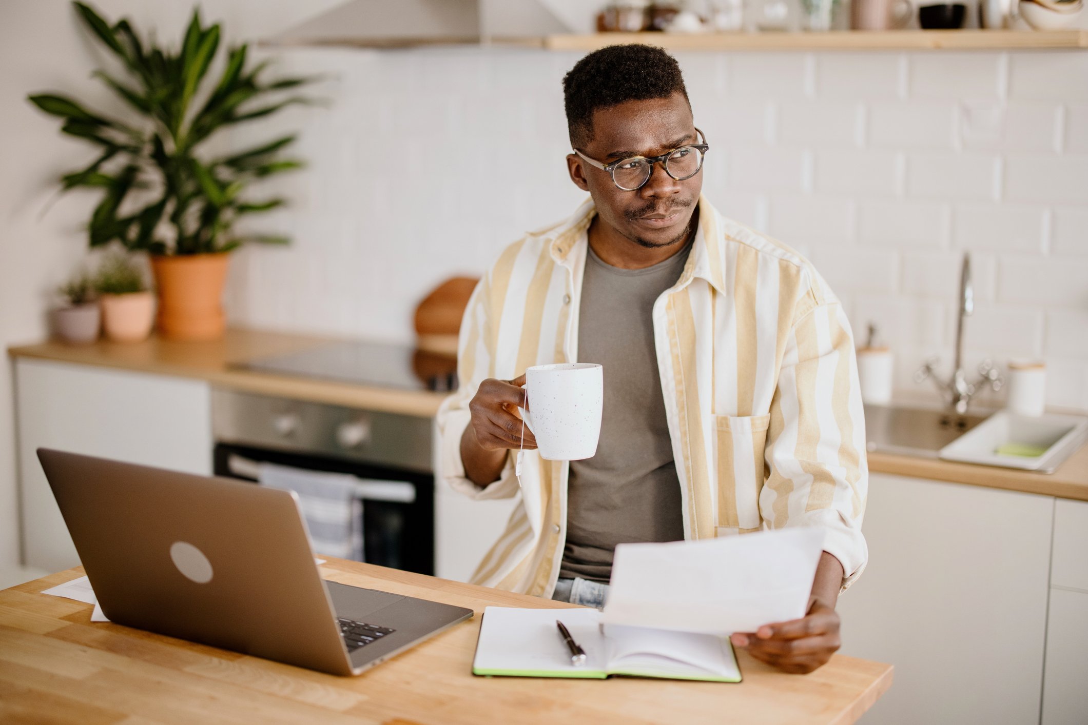 Man drinking coffee in his kitchen checks a laptop and a document.